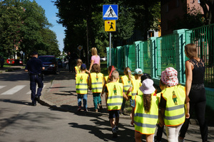 Policjant wraz z dziećmi w kamizelkach odblaskowych oraz opiekunkami idą w stronę oznakowanego przejścia dla pieszych.
