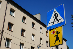 Widoczny na tle bloku mieszkalnego pionowy znak drogowy - przejście dla pieszych oraz tak zwana Agatka -  znak, który ostrzega o miejscu na drodze szczególnie uczęszczanym przez dzieci lub o bliskości takiego miejsca.