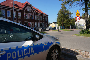 Na zdjęciu widoczny radiowóz oraz umundurowana policjantka obserwująca ruch drogowy na jezdni przed szkołą.