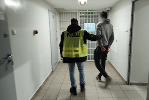 Na zdjęciu widoczny nieumundurowany policjant w kamizelce odblaskowej z napisem Policja, który prowadzi mężczyznę korytarzem. Mężczyzna na ręce trzymane z tyłu ma założone kajdanki.