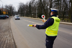 Na zdjęciu widoczny umundurowany policjant, który daje znak kierującemu samochodem, żeby zjechał na pobocze.