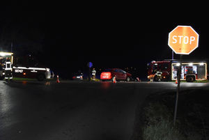 Na zdjęciu widoczne miejsce wypadku drogowego. Widoczne są tu dwa samochody osobowe oraz wozy strażackie i pracujący strażacy, a także znak Stop.
