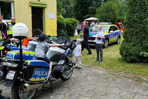 Na zdjęciu widoczne dziecko siedzące na motocyklu. Obok stoi policjant. Widoczne te4ż dzieci przyglądające się motocyklom. Za nimi stoi radiowóz.