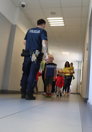 Na zdjęciu widoczny policjant oprowadzający korytarzem dzieci i opiekunów.