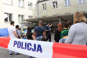 Na zdjęciu widoczny policjant oraz dzieci i opiekunowie na placu wewnętrznym zawierciańskiej jednostki Policji. Policjant wskazuje przygotowane do zwiedzania stoiska.