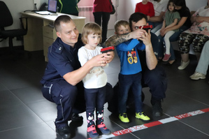 Na zdjęciu widoczna dziewczynka oraz chłopiec oraz dwaj policjanci, którzy im pomagają strzelać z broni ćwiczebnej na strzelnicy multimedialnej.