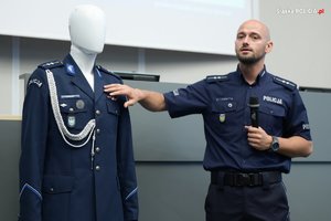 Zdjęcie przedstawia umundurowanego policjanta, który w lewej dłoni trzyma mikrofon a prawą wskazuje na mundur na manekinie.