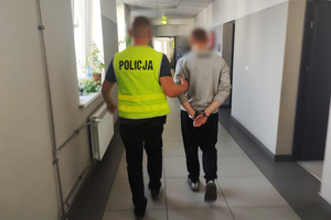 Na zdjęciu widoczny nieumundurowany policjant w kamizelce odblaskowej z napisem Policja, który prowadzi korytarzem zatrzymanego mężczyznę. Mężczyzna na rękach trzymanych z tyłu, ma założone kajdanki.