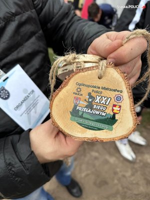 Zbliżenie na pamiątkowy medal Ogólnopolskich Mistrzostw Policji w XXI Biegu Przełajowym im. Grzegorza Załogi trzymany w dłoniach przedstawiciela organizatora.