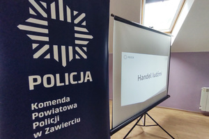 Na zdjęciu widoczny baner z policyjną gwiazdą i napisem Komenda Powiatowa Policji w Zawierciu oraz wyświetlony slajd prezentacji multimedialnej z napisem: Handel ludźmi.