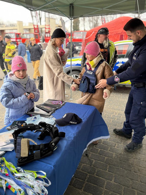 Na zdjęciu widoczny policjant przy stoisku profilaktycznym oraz dzieci i dorośli przy nim. Dziewczynka przymierza policyjną kamizelkę.