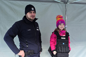 Na zdjęciu widoczny policjant oraz dziewczynka, która ma ubraną kamizelkę policyjną.