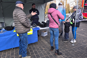 Na zdjęciu widoczne stoisko profilaktyczne, a także policjant i osoby stojące przy stoisku. Kobieta trzyma w rękach tarczę, a dziecko pałkę.