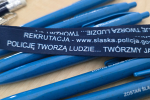 Na zdjęciu widoczne materiały promocyjne w postaci ołówków i smyczy. Na ołówkach widoczny napis: Zostań śląskim policjantem. Na smyczach widoczny napis: Rekrutacja - www.slaska. policja.gov.pl, Policję tworzą ludzie... Twórzmy ją razem.