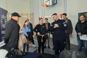 Na zdjęciu widoczny policjant, który prezentuje policyjny sprzęt uczestnikom spotkania. Jedna z kobiet ma na sobie policyjną kamizelkę.