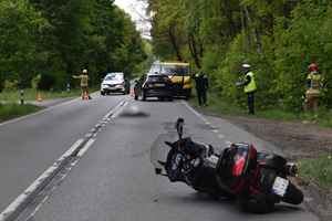 Na zdjęciu widoczny motocykl, który leży na jezdni. Nieco dalej na jezdni widoczne dwa samochody, a na poboczu laweta. Po prawej stronie widoczny policjant, strażak i idący poboczem mężczyzna. Na jezdni ruchem kieruje strażak.