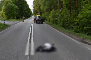 Na zdjęciu widoczna odzież i kask motocyklisty, leżące na jezdni. W dali widoczny samochód, który stoi na jezdni, a za nim laweta. Widoczny także strażak.