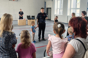Na zdjęciu widoczny stojący na sali gimnastycznej policjant, który trzyma za rękę dziewczynkę. Widoczne także stanowiska ze sprzętem policyjnym oraz dzieci z opiekunami.