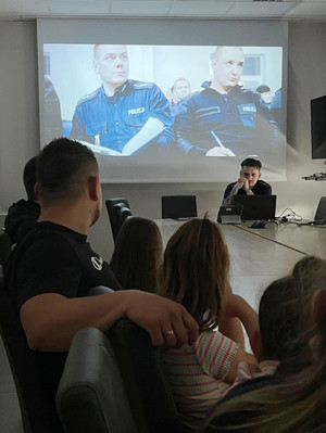 Na zdjęciu widoczne dzieci siedzące przy stole konferencyjnym i oglądające film o Policji. Widoczny także siedzący policjant.