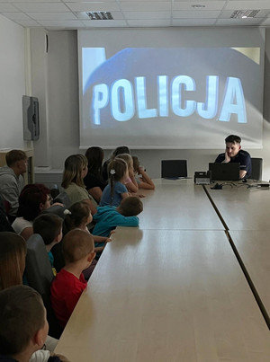 Na zdjęciu widoczne dzieci siedzące przy stole konferencyjnym i oglądające film o Policji. Widoczny także siedzący policjant.