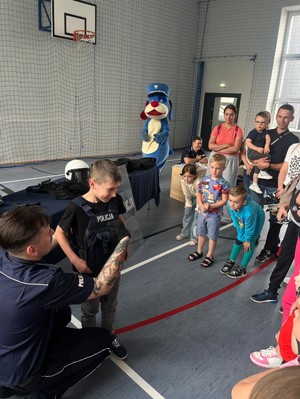 Na zdjęciu widoczni policjanci, dzieci, opiekunowie i Sznupek na sali gimnastycznej. Jeden z chłopców przymierza kamizelkę policyjną. Pomaga mu w tym policjant.
