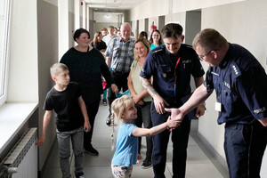Na zdjęciu widoczny policjant, który prowadzi korytarzem dzieci i opiekunów, a także dziewczynka, która podaje rękę innemu policjantowi.