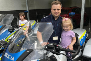 Na zdjęciu widoczny policjant i dziewczynka, która siedzi na motocyklu. W tle widoczna inna dziewczynka, która również siedzi na motocyklu.