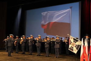 Na scenie widoczna orkiestra Komendy Wojewódzkiej Policji w Katowicach oraz poczet sztandarowy. W tle wyświetlona powiewająca flaga Polski.
