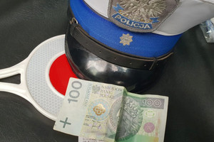 Na zdjęciu widoczna policyjna czapka, tarcza do zatrzymywania pojazdów, a także 100 złotowy banknot.