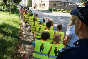 Na zdjęciu widoczne dzieci w kamizelkach odblaskowych, które idą chodnikiem z opiekunami, a także policjant.