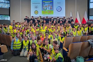 Na zdjęciu widoczne dzieci w kamizelkach odblaskowych oraz orkiestra Komendy Wojewódzkiej Policji w Katowicach.