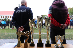 Na zdjęciu widoczne puchary, Pierwszy Zastępca Komendanta Powiatowego Policji w Zawierciu podinspektor Paweł Sobota oraz mężczyzna stojący obok. W tle widoczni zawodnicy turnieju.