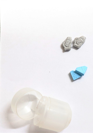 Na zdjęciu widoczne plastikowe opakowanie, a także dwie tabletki koloru szarego oraz dwie tabletki koloru niebieskiego.