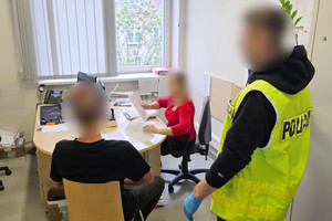 Na zdjęciu widoczny nieumundurowany policjant w kamizelce odblaskowej z napisem POLICJA, który stoi po prawej stronie. Widoczna także nieumundurowana policjantka, która siedzi przy biurku i przekłada dokumenty, a także zatrzymany mężczyzna, który siedzi przy biurku.
