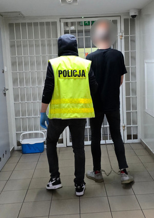 Na zdjęciu widoczny nieumundurowany policjant w kamizelce odblaskowej z napisem POLICJA, który prowadzi korytarzem zatrzymanego mężczyznę. Mężczyzna ma założone kajdanki zespolone.