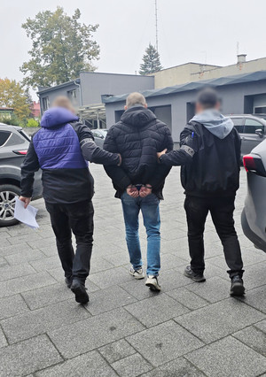 Na zdjęciu widoczni dwaj nieumundurowani policjanci z opaskami na ramionach z napisem POLICJA, którzy prowadzą zatrzymanego mężczyznę. Mężczyzna ma założone kajdanki na ręce trzymane z tyłu.