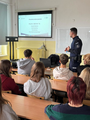 Na zdjęciu widoczny policjant, podczas prelekcji dla uczniów, którzy siedzą w ławkach.