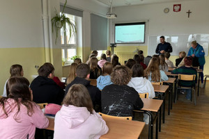 Na zdjęciu widoczny policjant, podczas prelekcji dla uczniów, którzy siedzą w ławkach.