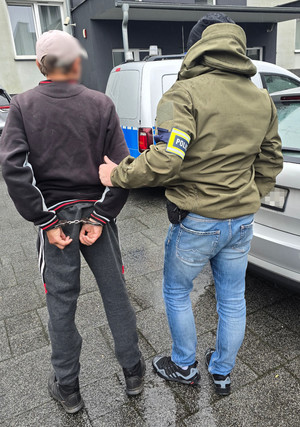 Na zdjęciu widoczny nieumundurowany policjant, który prowadzi zatrzymanego mężczyznę. Mężczyzna ma założone kajdanki na ręce trzymane z tyłu.