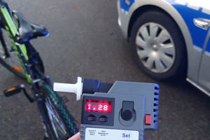 Na zdjęciu widoczne urządzenie do badania stanu trzeźwości z wyświetlonym wynikiem 1,28. W tle widoczny częściowo radiowóz oraz rower.