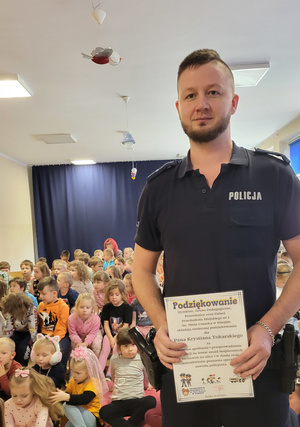 Na zdjęciu widoczny policjant, który trzyma pamiątkowy dyplom. Za nim widoczne siedzące dzieci.
