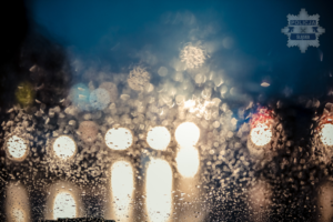 Na zdjęciu widoczne przez mokrą szybę samochody z włączonymi światłami.