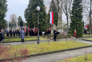 Na zdjęciu widoczny Pomnik Niepodległości na cmentarzu oraz stojąca przed nim warta, policjant i mężczyzna przy mikrofonie.