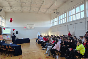 Na zdjęciu widoczny policjant prowadzący spotkanie z młodzieżą, zgromadzoną na sali gimnastycznej.