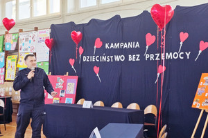 Na zdjęciu widoczny policjant stojący na tle kotary z napisem: Kampania Dzieciństwo bez Przemocy.