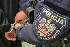Na zdjęciu widoczny częściowo policjant, który zakłada kajdanki mężczyźnie na ręce trzymane z tyłu.