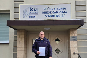 Na zdjęciu widoczny umundurowany policjant trzymający w ręku plakaty, przed budynkiem Spółdzielni Mieszkaniowej "Zawiercie".
