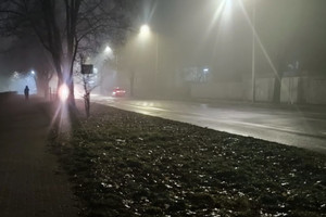 Na zdjęciu widoczna jezdnia nocą, mgła, jadący pojazd i pieszy idący chodnikiem.