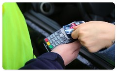 Na zdjęciu widoczny terminal płatniczy trzymany w ręku przez policjanta, a także kierujący, który zbliża kartę płatniczą do terminala.