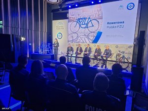 Na zdjęciu widoczne osoby siedzące na scenie oraz publiczność.